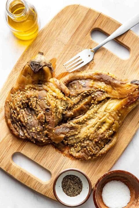 Roasted eggplant on a wooden cutting board, with a fork, a jar of oil, and small bowls of salt and pepper nearby.