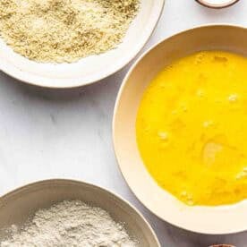 Three bowls with flour, beaten eggs, and breadcrumbs, alongside small bowls of salt and pepper, arranged on a white surface.