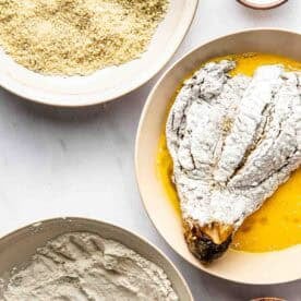 A breading station with bowls of flour, beaten eggs, breadcrumbs, black pepper, salt, and an eggplant coated in flour ready for dipping in egg.