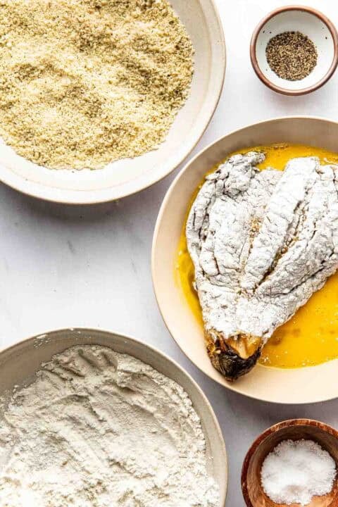 A breading station with bowls of flour, beaten eggs, breadcrumbs, black pepper, salt, and an eggplant coated in flour ready for dipping in egg.