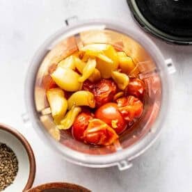 Overhead view of a blender cup filled with roasted tomatoes, onions, and garlic, surrounded by small bowls of salt and pepper on a white surface.