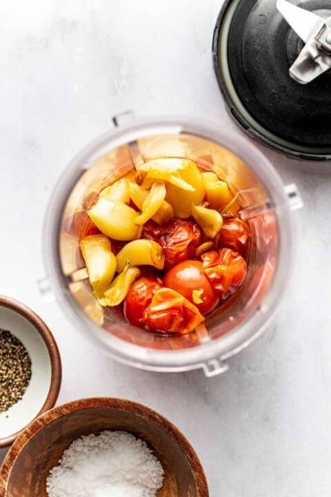Overhead view of a blender cup filled with roasted tomatoes, onions, and garlic, surrounded by small bowls of salt and pepper on a white surface.