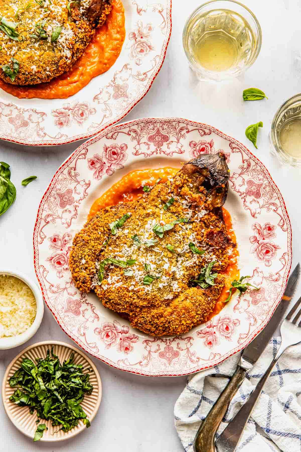 A breaded eggplant sits on a red-and-white plate over tomato sauce, garnished with herbs and grated cheese, beside small bowls of cheese and chopped herbs, and glasses of white wine.