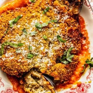 A breaded and baked eggplant half topped with grated cheese and fresh basil, served on tomato sauce on a floral plate, with a fork cutting into it.