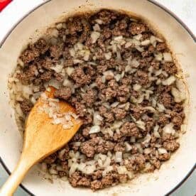 Ground beef and chopped onions are being cooked in a large pot, with a wooden spoon resting inside.