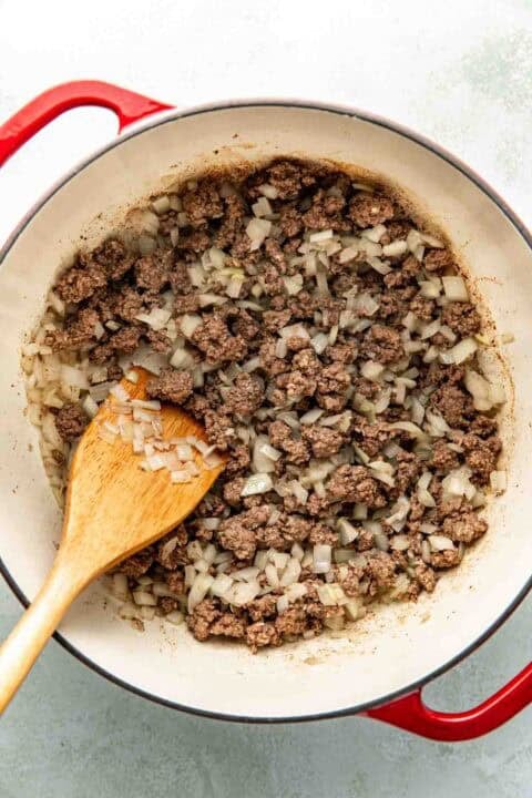 Ground beef and chopped onions are being cooked in a large pot, with a wooden spoon resting inside.