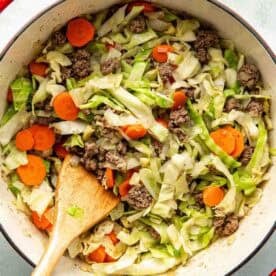 A red pot filled with cooked ground beef, sliced carrots, and shredded cabbage, with a wooden spoon resting inside.