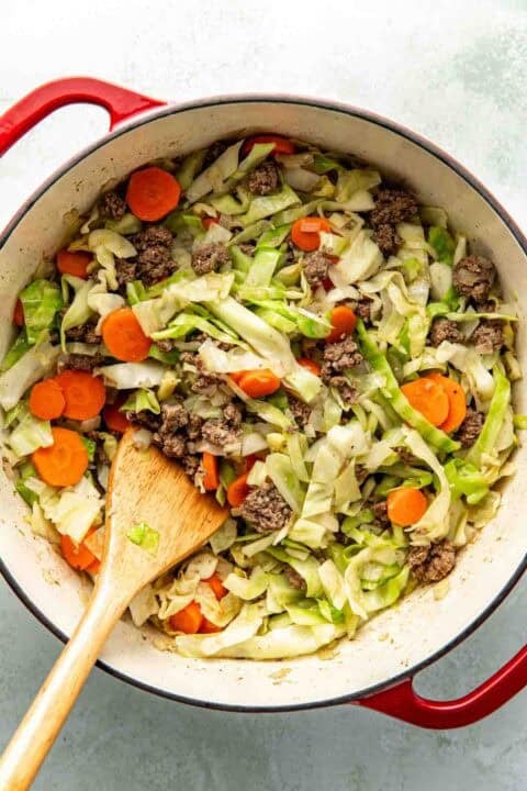 A red pot filled with cooked ground beef, sliced carrots, and shredded cabbage, with a wooden spoon resting inside.