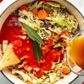 A pot of soup with ground beef, cabbage, carrots, tomato sauce, bay leaves, and broth being poured in, with a wooden spoon in the pot.