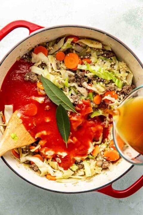 A pot of soup with ground beef, cabbage, carrots, tomato sauce, bay leaves, and broth being poured in, with a wooden spoon in the pot.