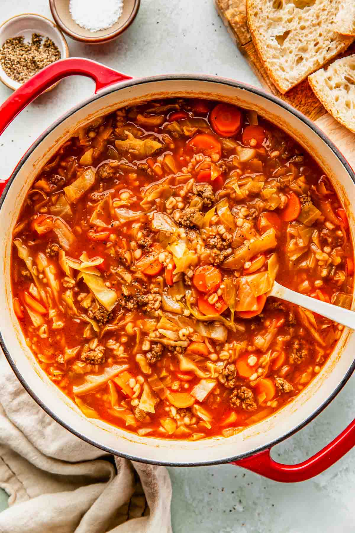 A pot of cabbage soup with ground beef, carrots, and tomato broth sits on a counter with a wooden spoon, bread slices, and small bowls of salt and pepper nearby.