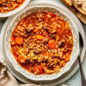 A bowl of beef and barley soup with carrots, cabbage, ground meat, and barley, served with slices of bread on the side.