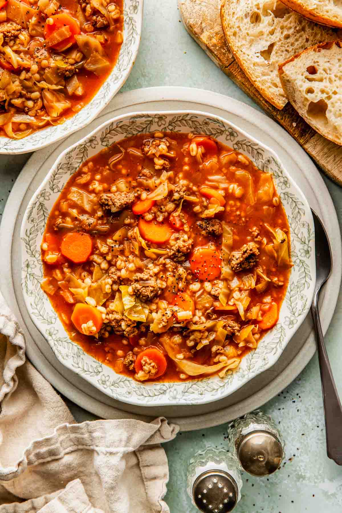 A bowl of beef and barley soup with carrots, cabbage, ground meat, and barley, served with slices of bread on the side.