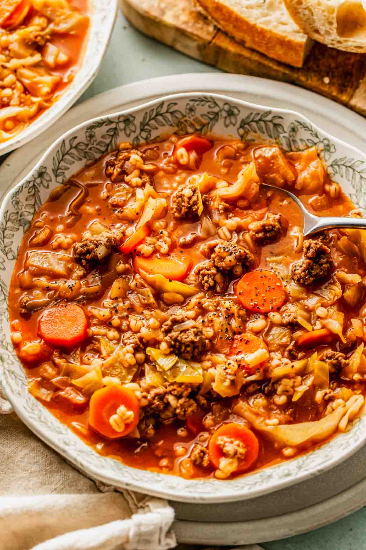 A bowl of vegetable and ground beef soup with carrots, cabbage, tomatoes, and barley, served with a spoon.