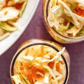 Two glass jars filled with shredded cabbage and carrots, with more of the vegetable mixture visible on a plate in the background.