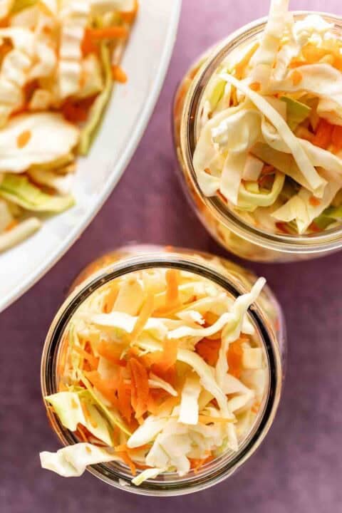 Two glass jars filled with shredded cabbage and carrots, with more of the vegetable mixture visible on a plate in the background.