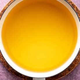 A white saucepan filled with clear yellow brining liquid, placed on a round cork pad against a purple textured background.
