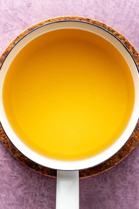 A white saucepan filled with clear yellow brining liquid, placed on a round cork pad against a purple textured background.