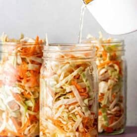 Three glass jars filled with shredded cabbage and carrots, with liquid being poured into one jar from a white container.