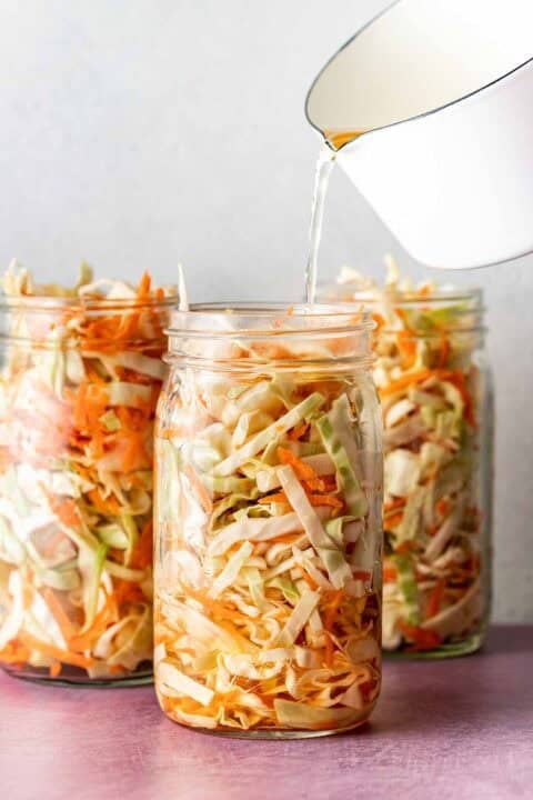 Three glass jars filled with shredded cabbage and carrots, with liquid being poured into one jar from a white container.