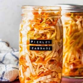 A glass jar filled with pickled cabbage sits on a pink surface, labeled Pickled Cabbage; another jar and a fork are beside it.