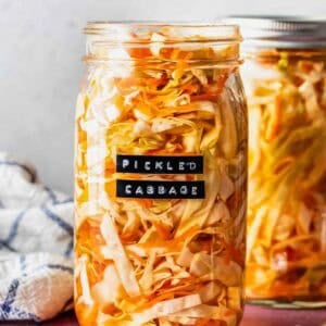 A glass jar filled with pickled cabbage sits on a pink surface, labeled Pickled Cabbage; another jar and a fork are beside it.
