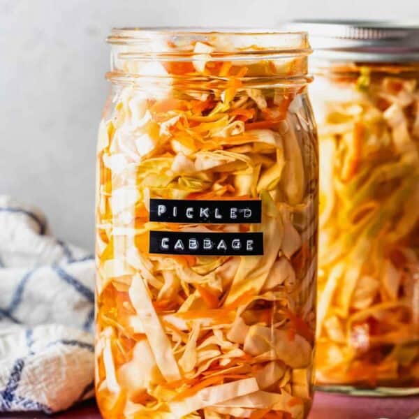 A glass jar filled with pickled cabbage sits on a pink surface, labeled Pickled Cabbage; another jar and a fork are beside it.