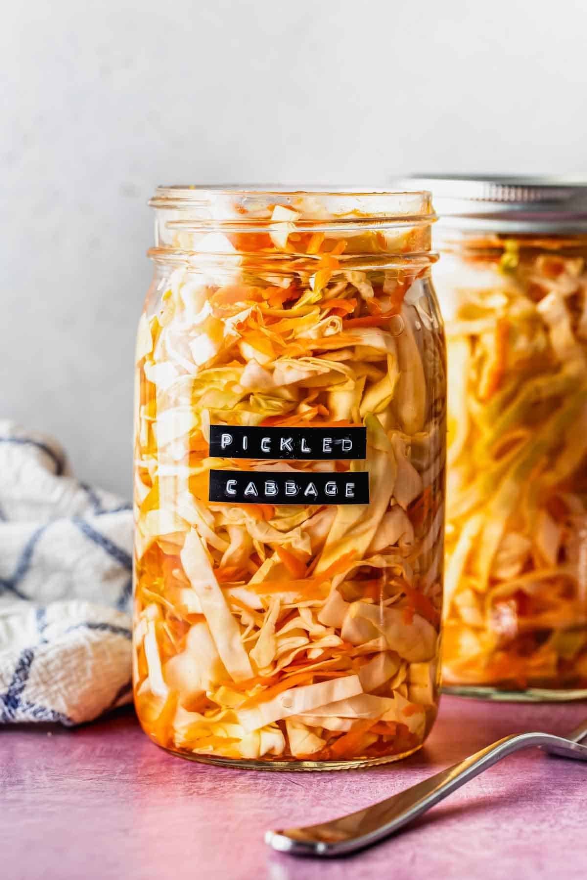A glass jar filled with pickled cabbage sits on a pink surface, labeled Pickled Cabbage; another jar and a fork are beside it.