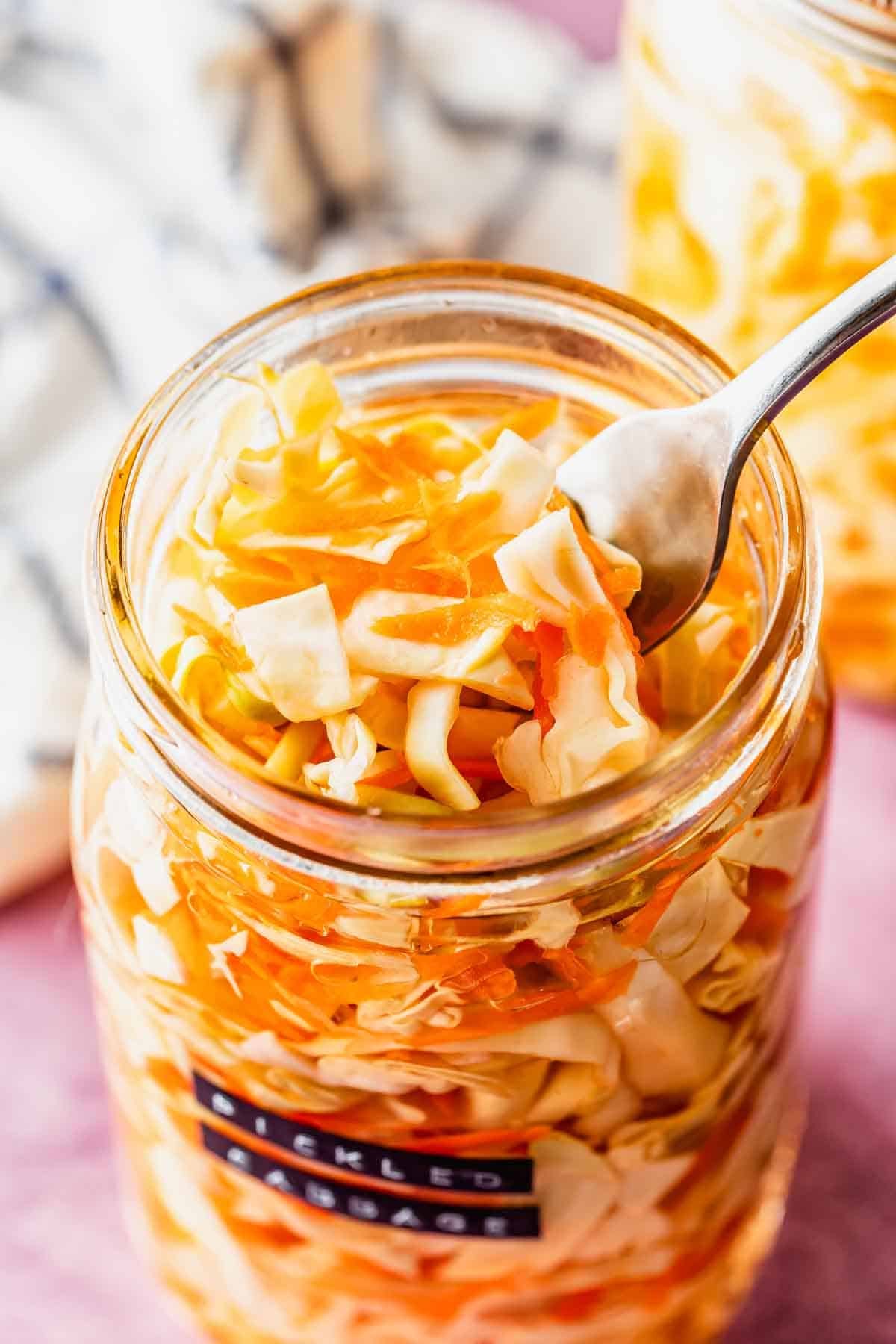 A spoon in a jar of pickled cabbage and carrots, with a striped cloth and another jar in the background.