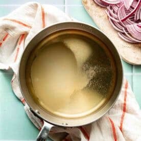 A saucepan filled with liquid sits on a striped kitchen towel next to a cutting board with sliced red onions on a light blue countertop.