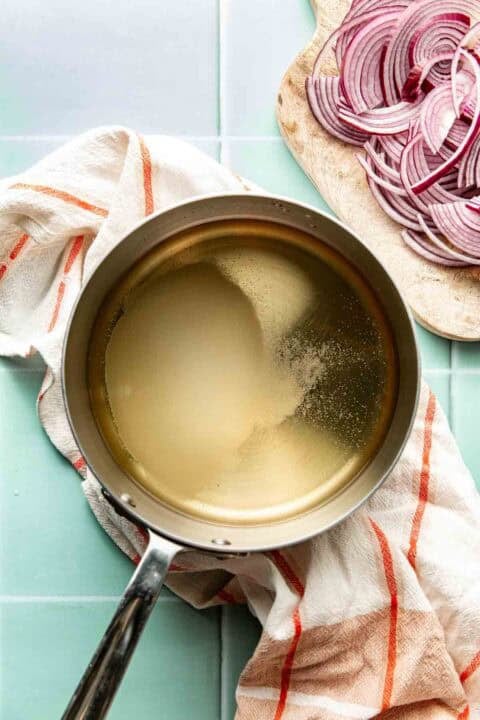 A saucepan filled with liquid sits on a striped kitchen towel next to a cutting board with sliced red onions on a light blue countertop.