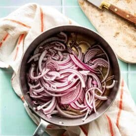 A stainless steel pan filled with thinly sliced red onions sits on a striped kitchen towel, next to a cutting board and knife.