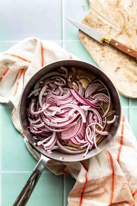 A stainless steel pan filled with thinly sliced red onions sits on a striped kitchen towel, next to a cutting board and knife.