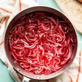 A saucepan filled with sliced red onions soaking in a red liquid, placed on a light towel next to a wooden cutting board and a knife.