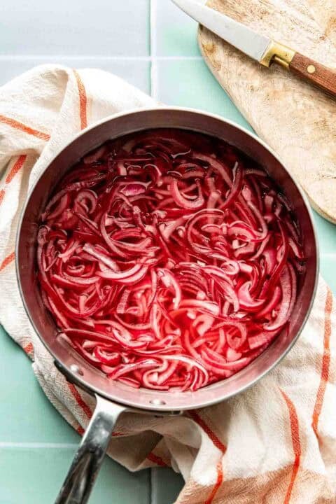 A saucepan filled with sliced red onions soaking in a red liquid, placed on a light towel next to a wooden cutting board and a knife.