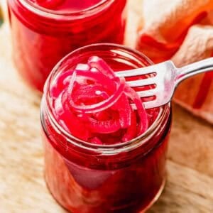 A fork holds sliced pickled red onions above an open glass jar, with another jar and a cloth visible in the background on a wooden surface.