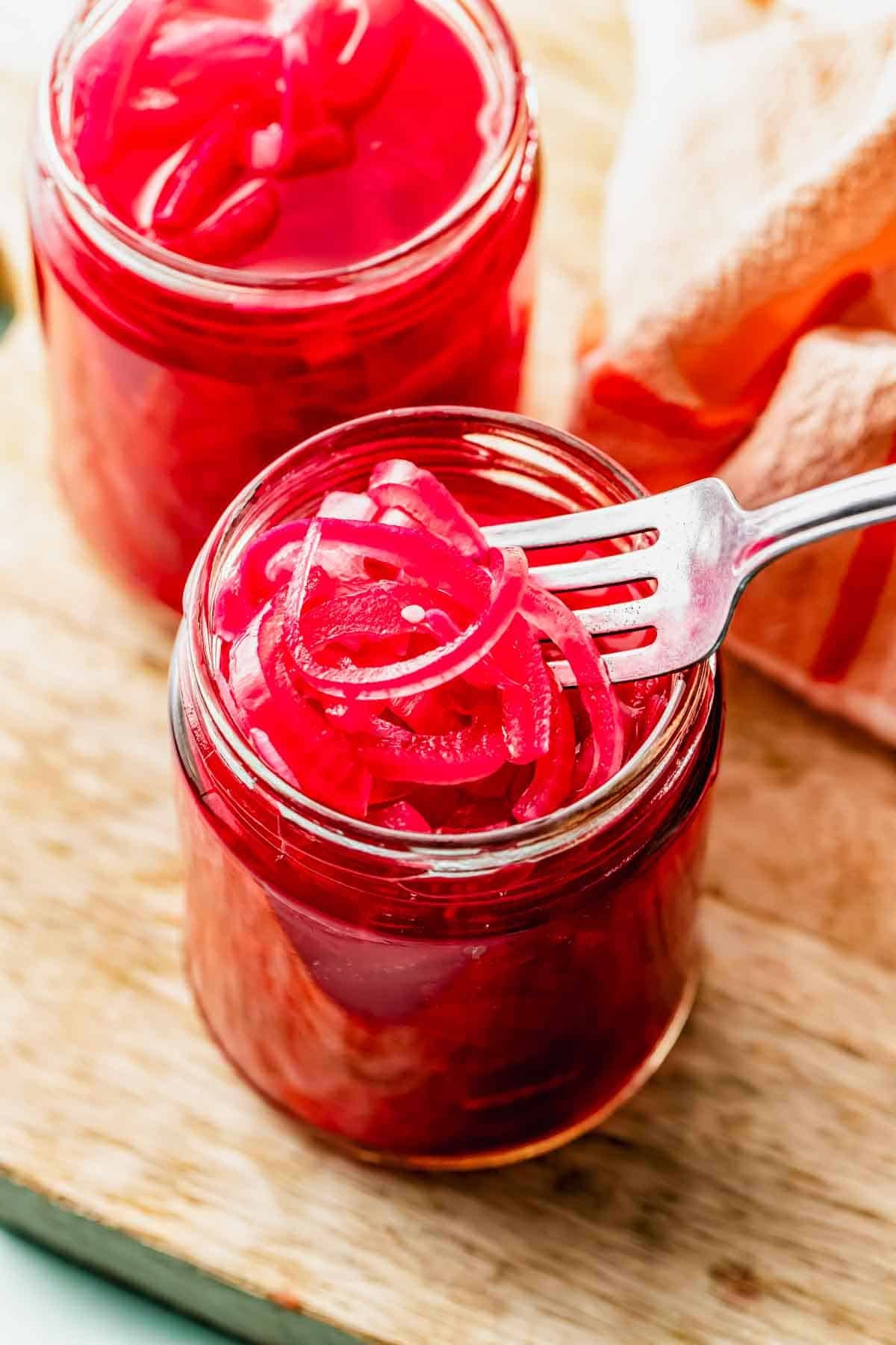 A fork holds sliced pickled red onions above an open glass jar, with another jar and a cloth visible in the background on a wooden surface.