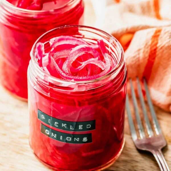 Two jars of pickled red onions sit on a wooden surface next to a fork, with a labeled jar in the foreground and a striped cloth in the background.