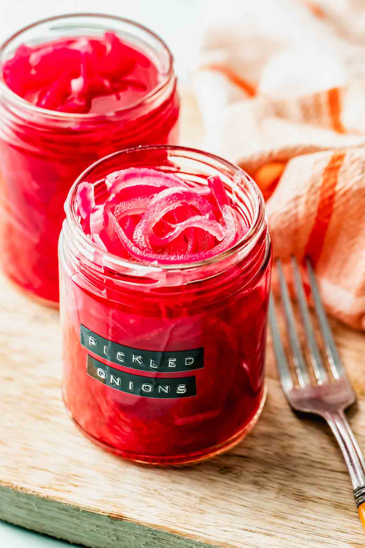 Two jars of pickled red onions sit on a wooden surface next to a fork, with a labeled jar in the foreground and a striped cloth in the background.
