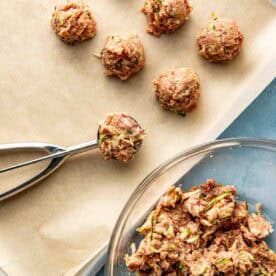 A baking sheet lined with parchment holds raw meatball mixtures, with a cookie scoop and a glass bowl of the meat mixture nearby.