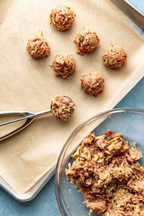 A baking sheet lined with parchment holds raw meatball mixtures, with a cookie scoop and a glass bowl of the meat mixture nearby.