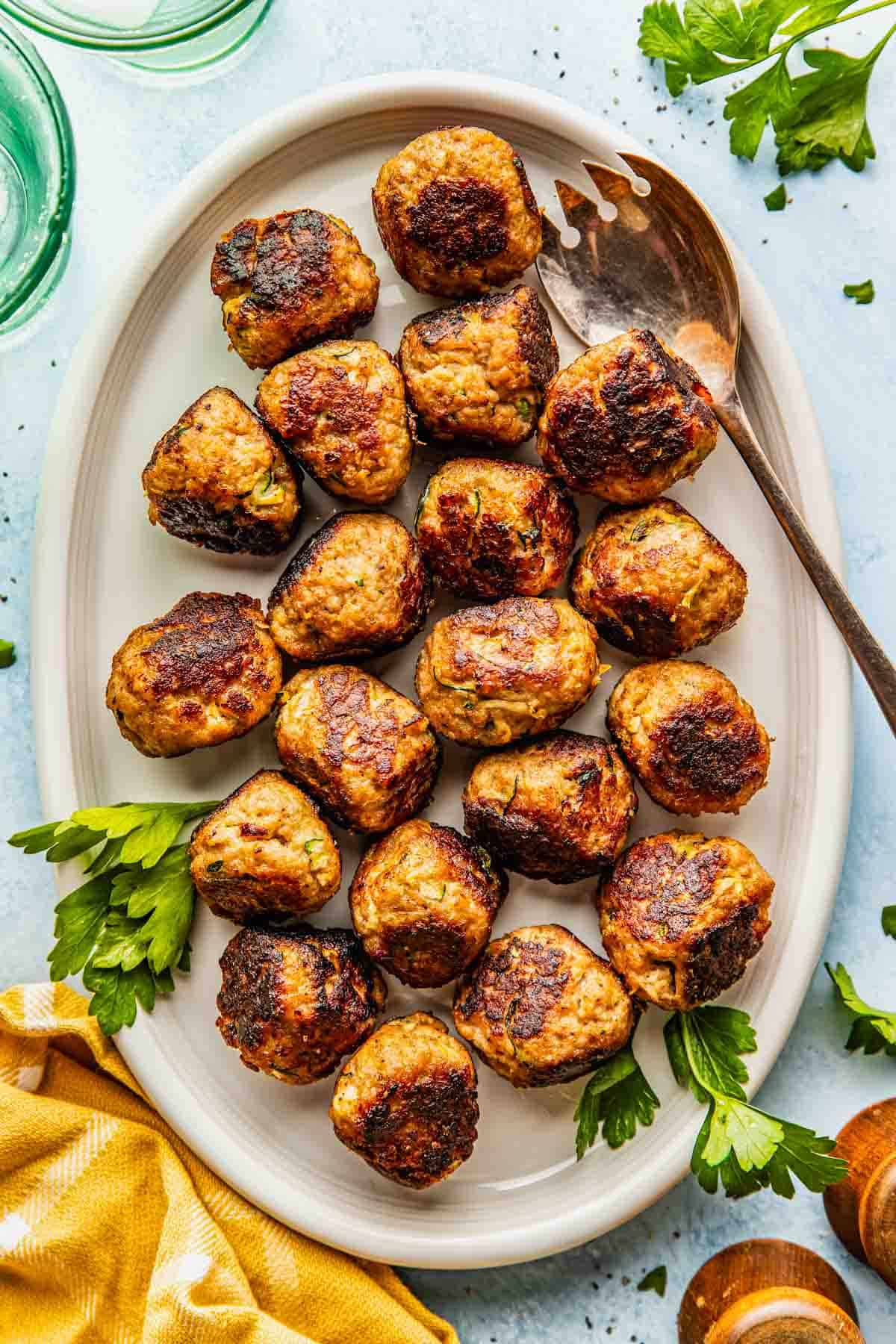 A white oval plate with twenty browned meatballs, garnished with parsley, and a metal serving spoon on the side.