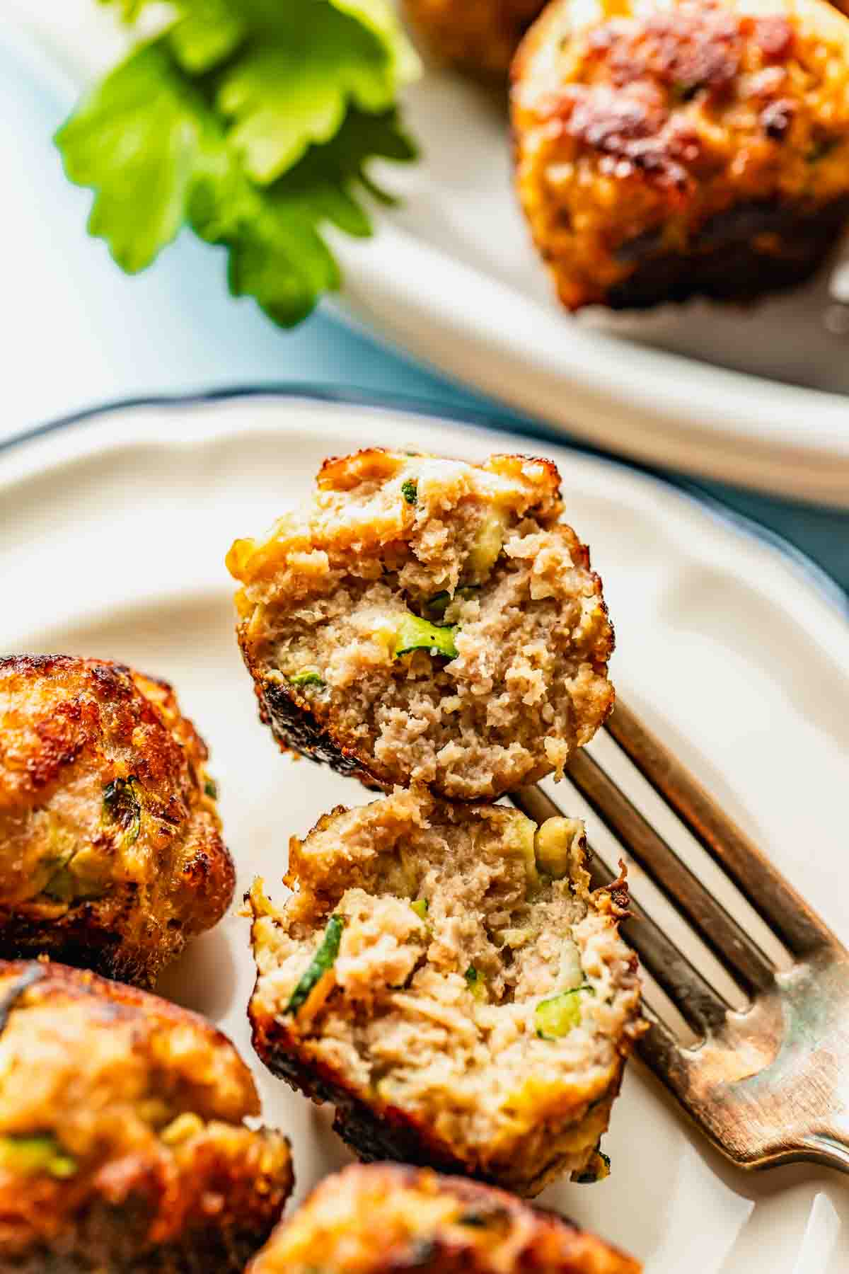 A close-up of a plate with meatballs, one cut open with a fork to show the inside texture and ingredients like herbs and vegetables. Parsley garnish is visible in the background.
