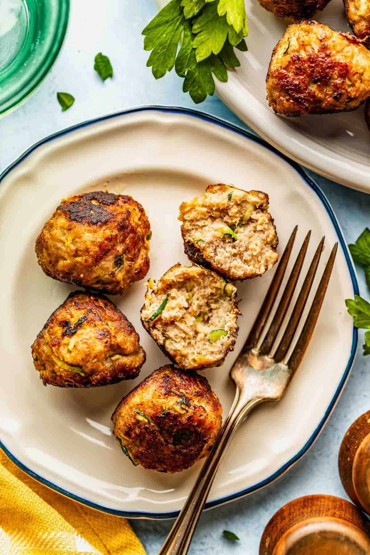 A plate with several cooked meatballs, some whole and one cut in half to show the inside, with a fork placed beside them and parsley garnish nearby.