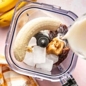 A blender contains ice cubes, a banana, dates, and peanut butter. Milk is being poured in. Bananas and a striped towel are nearby on a pink surface.