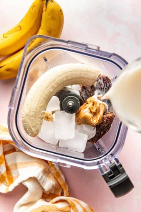 A blender contains ice cubes, a banana, dates, and peanut butter. Milk is being poured in. Bananas and a striped towel are nearby on a pink surface.