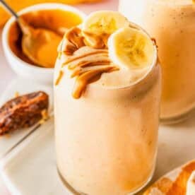 A glass of banana smoothie topped with banana slices and peanut butter, with a bowl of peanut butter, dates, and a banana in the background.
