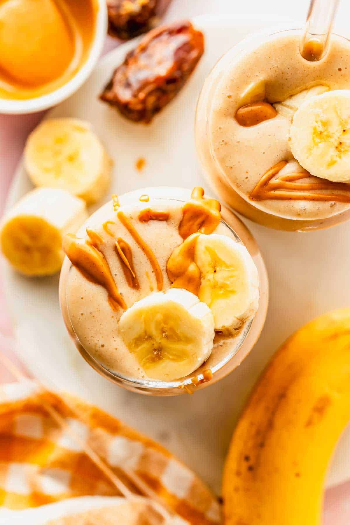 Zwei Tassen Bananen-Smoothie, garniert mit Bananenscheiben und beträufelt mit Erdnussbutter, umgeben von Bananen, Datteln und einer Schüssel Erdnussbutter auf einer weißen Oberfläche.
