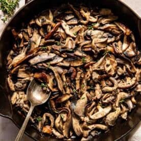 A cast iron skillet filled with sautéed mixed mushrooms, garnished with fresh herbs, with a spoon resting inside.
