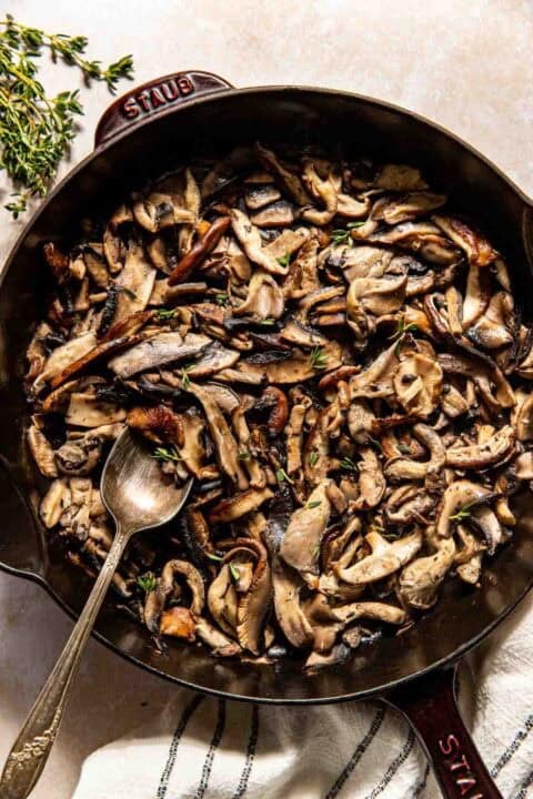 A cast iron skillet filled with sautéed mixed mushrooms, garnished with fresh herbs, with a spoon resting inside.